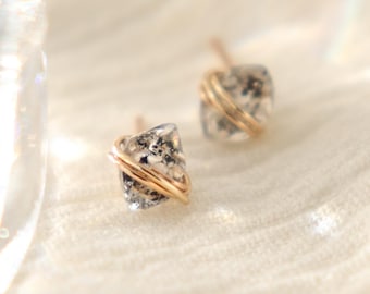 May include: A pair of small, square stud earrings. Each earring features a clear, speckled stone wrapped with thin, gold-colored wire. The earrings are set against a soft, white background, with a blurred glass object in the upper left corner.