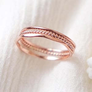 Può includere: Un anello in oro rosa con un design strutturato. L'anello presenta un motivo a corda intrecciata al centro, con un delicato motivo a perline sulla fascia inferiore. L'anello è posto su uno sfondo bianco.