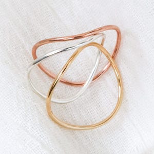 May include: Three minimalist rings in gold, silver, and rose gold tones are arranged on a white fabric surface. Each ring has a unique, organic shape, creating a modern and elegant jewelry set. The rings are simple and delicate.