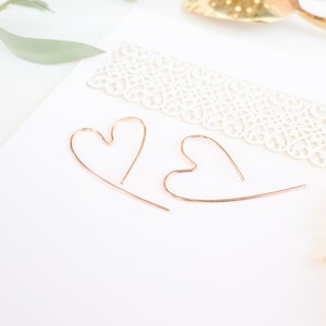 Può includere: Due delicati orecchini a forma di cuore, in oro rosa, su una superficie bianca. Gli orecchini sono a filo e di design minimalista. Sono visibili anche un bordo in pizzo decorativo e un cucchiaio dorato.