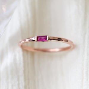 May include: A rose gold ring with a hammered band and a rectangular, magenta-colored gemstone. The ring's design is simple and elegant, showcasing the vibrant color of the gemstone. The ring is a delicate piece of jewelry.
