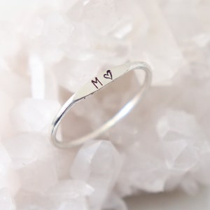 Dainty, sterling silver initial ring with the letter M and a heart stamp sits atop a sparkling white crystal background.  The ring is also available in rose gold filled and gold filled.