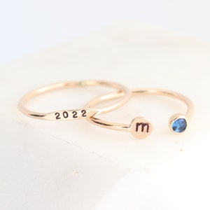 May include: Two gold rings, one engraved with the year 2022 and the other with the letter "m" and a small blue gemstone.