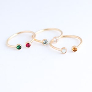 Three gold cuff rings are seen from a top view lying next to each other with glistening gemstones of various colours in each end. Rings are lying on an off-white background.