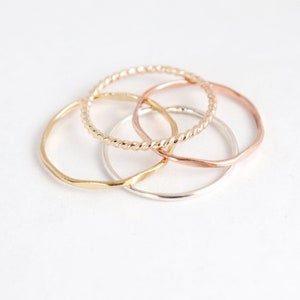 Four mixed metal gold, silver and rose rings sitting on a light background in a pile, shown from above. Ring textures can be seen up close: twisted rope, hammered, and smooth in different metals.