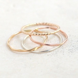 Close up view of four stackable rings in yellow gold, rose gold and sterling silver. The rings are in a pile on a light background with slight texture. The rings are stacked by smooth, hammered and twisted rope styles.
