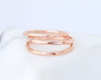 May include: Three delicate rose gold stacking rings. The top ring has a textured finish, the middle ring is smooth, and the bottom ring has a hammered finish.