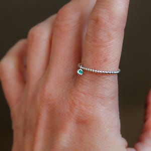 K&ouml;nnte beinhalten: Ein silberner Ring mit einem gedrehten Band und einem kleinen, runden, teal-farbenen Edelstein. Der Ring ist zart und schlicht, mit einem minimalistischen Design. Der Edelstein ist in einer L&uuml;nettenfassung gefasst.