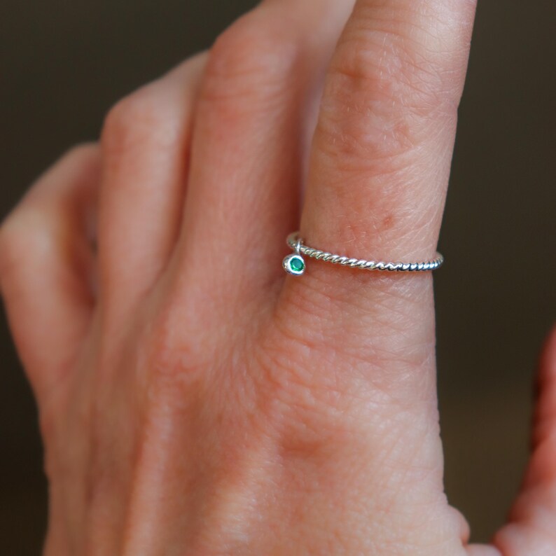 K&ouml;nnte beinhalten: Ein silberner Ring mit einem gedrehten Band und einem kleinen, runden, teal-farbenen Edelstein. Der Ring ist zart und schlicht, mit einem minimalistischen Design. Der Edelstein ist in einer L&uuml;nettenfassung gefasst.