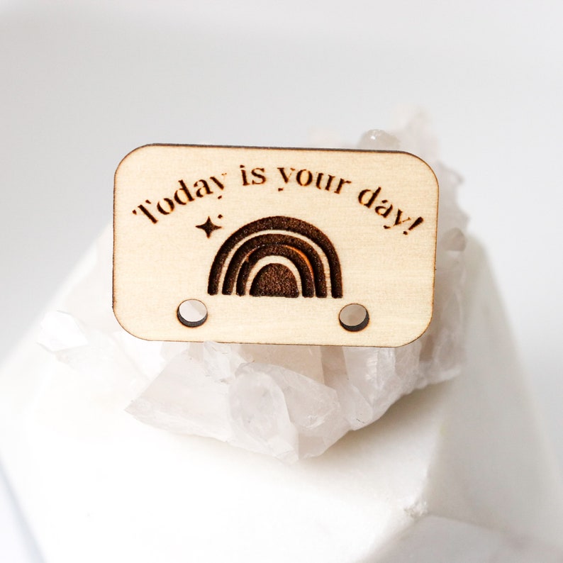May include: A rectangular wooden tag with the phrase "Today is your day!" and a rainbow design. The tag has two small holes and a star graphic. The wood has a light, natural color.