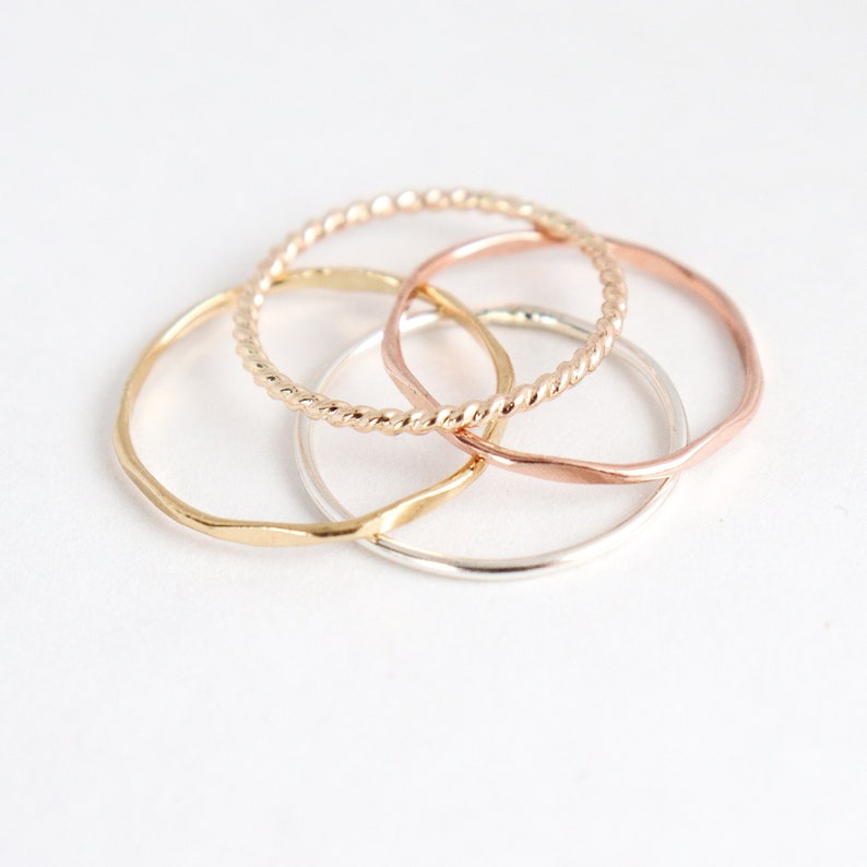 Four mixed metal gold, silver and rose rings sitting on a light background in a pile, shown from above. Ring textures can be seen up close: twisted rope, hammered, and smooth in different metals.