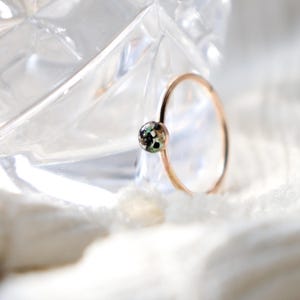 May include: A thin rose gold ring featuring a small, circular setting. The setting contains a speckled inlay with flecks of black, white, and green colors. The band is smooth and minimalist, showcasing a delicate design with a polished finish.