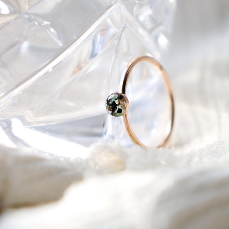 May include: A thin rose gold ring featuring a small, circular setting. The setting contains a speckled inlay with flecks of black, white, and green colors. The band is smooth and minimalist, showcasing a delicate design with a polished finish.