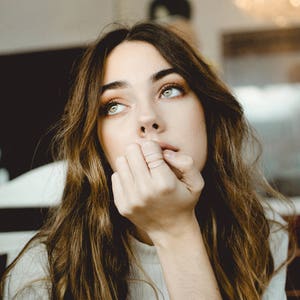 A woman with dramatic, light green eyes rests her head on her hand, as she sits in a cafe. Slim stackable mixed metal textured rings are featured on her fingers that are resting against her chin.