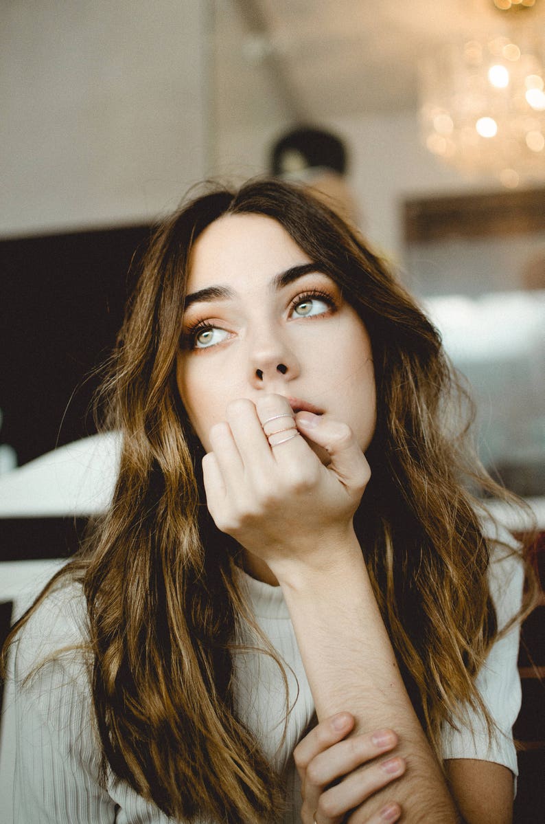 A woman with dramatic, light green eyes rests her head on her hand, as she sits in a cafe. Slim stackable mixed metal textured rings are featured on her fingers that are resting against her chin.