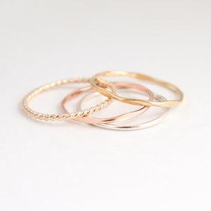 Four mixed metal gold, silver and rose rings sitting on a light background in a pile, shown from above. Ring textures can be seen up close: twisted rope, hammered, and smooth in different metals.