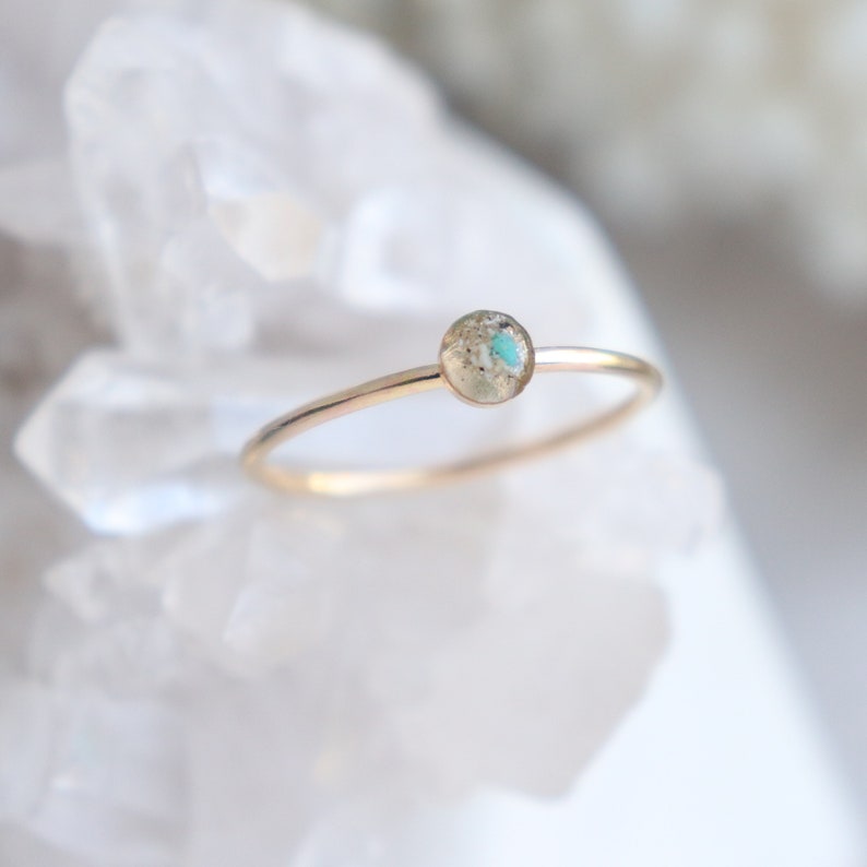May include: A thin gold band ring featuring a small, circular setting. The center of the setting contains a textured, neutral-toned inlay with a distinct, bright turquoise blue speck embedded within the surface of the circular focal point.