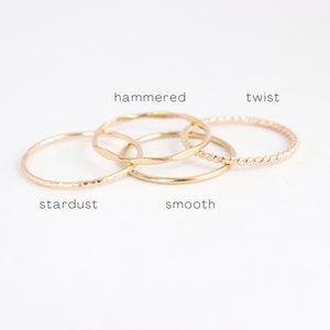 Four slim, stackable gold bands are shown neatly arranged in a pile. Text indicates the different style options featuring stardust, hammered, smooth and twist band options.