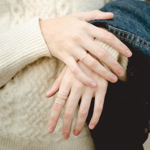 Four slim, stackable, textured metal ring bands are featured in a close up on a woman's hands. The hands are crossed over the front of her cream colored sweater. There is a denim jacket draped over one of her arms.