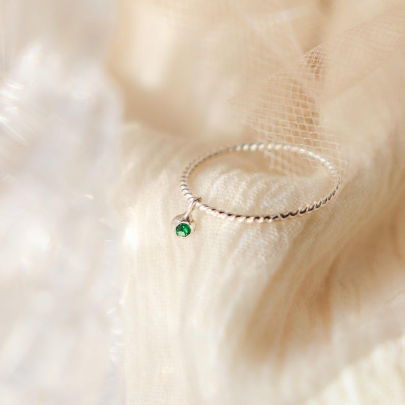 K&ouml;nnte beinhalten: Ein silberner Ring mit einem gedrehten Band und einem kleinen, h&auml;ngenden smaragdgr&uuml;nen Edelstein. Der Ring ist vor einem weichen, cremefarbenen Stoff platziert, wodurch eine zarte und elegante &Auml;sthetik entsteht. Das Design des Rings ist schlicht, aber auff&auml;llig.