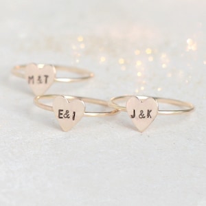 May include: Three gold-toned rings, each featuring a flat heart-shaped charm. The charms are stamped with black lettering: one reads M&T, another E&I, and the third J&K. The rings have thin, smooth bands and a polished metallic finish throughout.