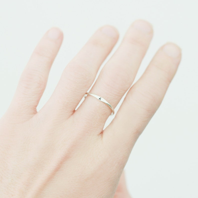 Slim stackable silver letter ring shown on the middle finger of a woman's hand to represent the size, color and shape detail of the dainty ring. Light and airy background.