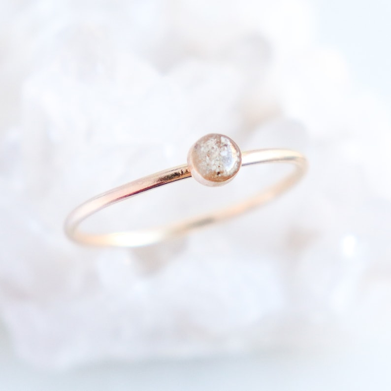 Hand made memorial cremation ring features ashes suspended in resin as a keepsake ring for individuals experiencing the loss of a loved one, human or pet. Gold ring is on a white, blurred background.