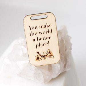 May include: A light-colored wooden tag with a rectangular shape and a small hole at the top. The tag features the phrase "You make the world a better place!" and a floral design, all in a dark brown color.