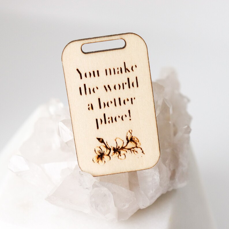 May include: A light-colored wooden tag with a rectangular shape and a small hole at the top. The tag features the phrase "You make the world a better place!" and a floral design, all in a dark brown color.