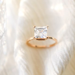 May include: A gold-colored ring with a large, square, clear gemstone. The ring is set on a white, textured surface. The gemstone is the focal point, reflecting light and adding sparkle.