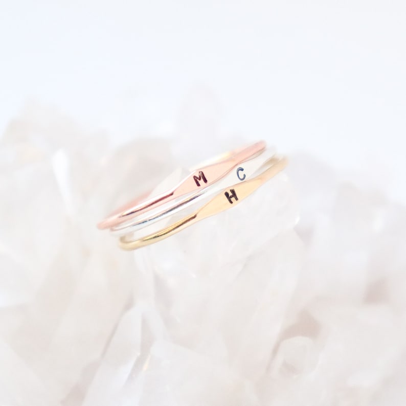 Three slim 1 mm stackable initial ring bands are stacked on a light white, crystal textured background that is slightly blurred. Sharp focus on the ring stack showing gold, silver and rose rings with initials H, C, M, respectively.