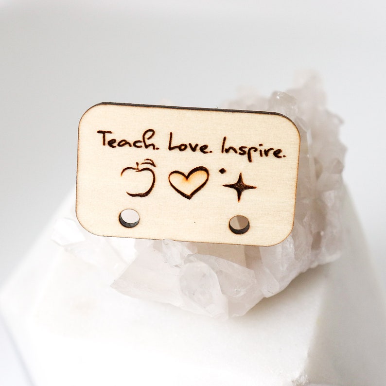 May include: A rectangular wooden tag with the words "Teach. Love. Inspire." etched on it. The tag features an apple, heart, and star symbol. Two small holes are at the bottom. The tag is light brown and sits on a white, textured surface.