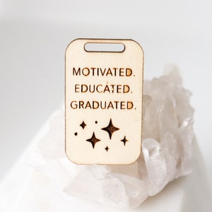 May include: A light-colored wooden tag with a rectangular shape and a hole at the top. The tag is engraved with the words "MOTIVATED. EDUCATED. GRADUATED." and four starburst designs. The tag is resting on a white, crystalline surface.