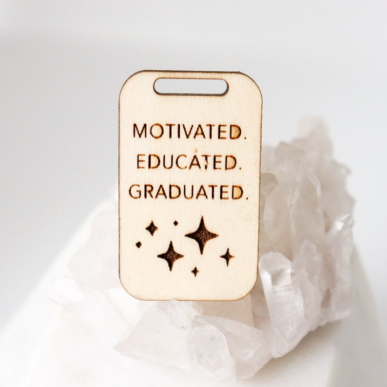 May include: A light-colored wooden tag with a rectangular shape and a hole at the top. The tag is engraved with the words "MOTIVATED. EDUCATED. GRADUATED." and four starburst designs. The tag is resting on a white, crystalline surface.