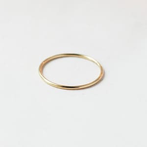 One single slim 1 mm rounded finger ring band in 14k yellow gold fill sits on a light off white background.