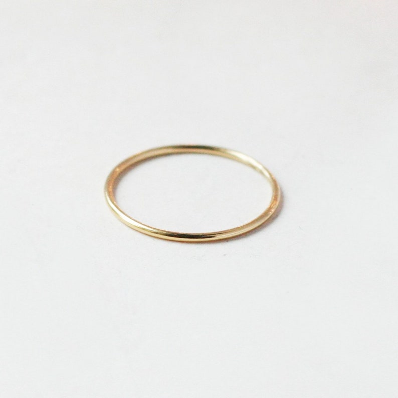 One single slim 1 mm rounded finger ring band in 14k yellow gold fill sits on a light off white background.