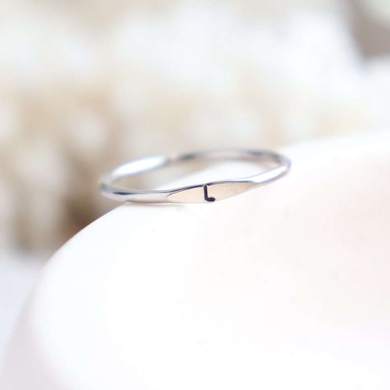 Smooth, round platinum stackable band with a small flat signet portion featuring the letter L hand stamped on the ring. The ring sits on the side of a display dish with blurred light textured background.