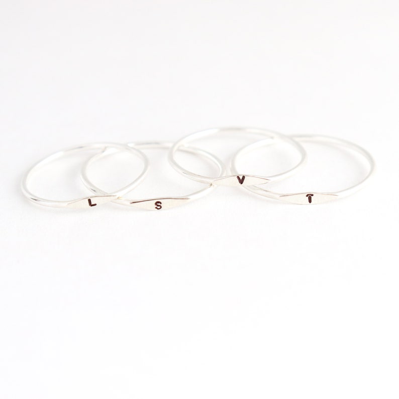 Four 1 mm handmade initial ring bands in sterling silver are neatly arranged in an overlapping stack featuring tiny letters L, S, V and T respectively from left to right. The rings sit on a very light off white background with tight focus on rings.