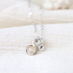 May include: A silver necklace with a small, round pendant featuring a delicate shell design. The pendant is attached to a silver chain with a small charm that reads "F".