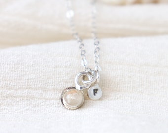 May include: A silver necklace with a small, round pendant featuring a delicate shell design. The pendant is attached to a silver chain with a small charm that reads "F".