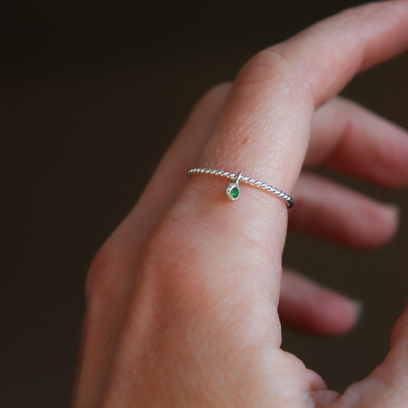 K&ouml;nnte beinhalten: Ein silberner Ring mit einem gedrehten Band und einem kleinen, h&auml;ngenden gr&uuml;nen Edelstein. Der Ring ist zart und minimalistisch und hat einen einzelnen, runden, smaragdgr&uuml;nen Stein. Der Ring wird an einem Finger getragen.