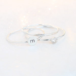 May include: Three silver rings stacked together. The top ring has a small clear stone, the middle ring is plain, and the bottom ring has the letter "M" stamped on it.