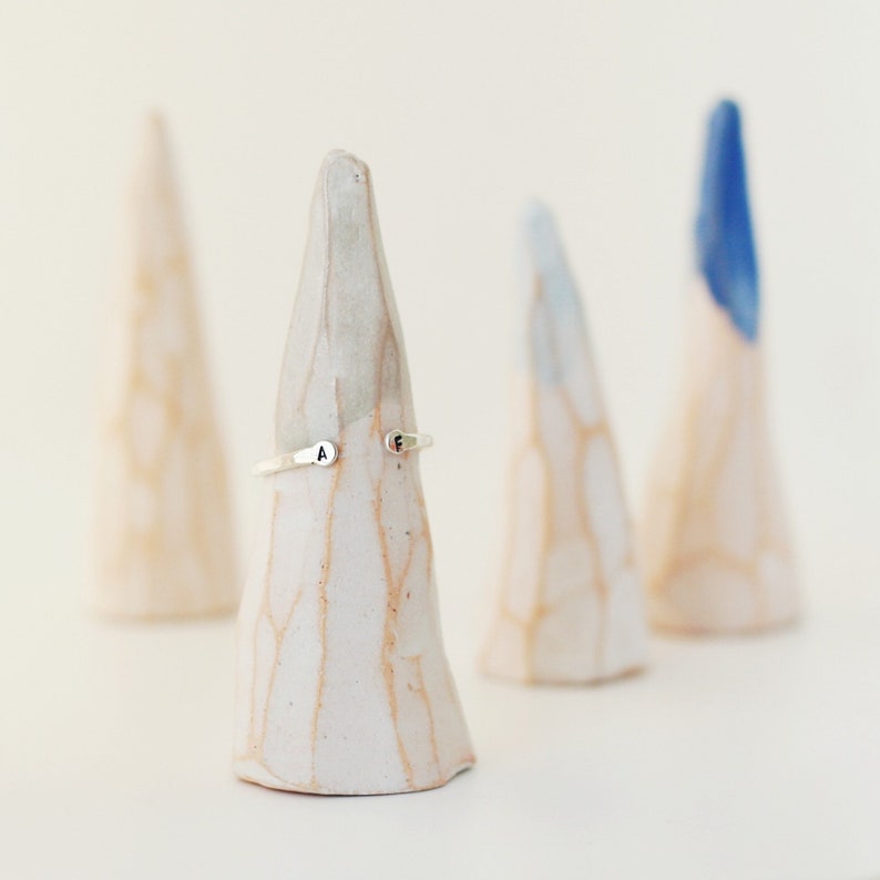 May include: A silver ring with the letters "A" and "F" engraved on it, displayed on a white ceramic cone. Three other ceramic cones are in the background, one with a blue tip. The ring is a simple, elegant design.