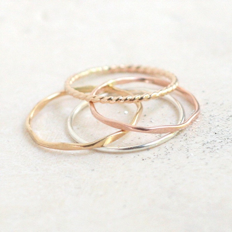 Close up view of four stackable rings in yellow gold, rose gold and sterling silver. The rings are in a pile on a light background with slight texture. The rings are stacked by smooth, hammered and twisted rope styles.