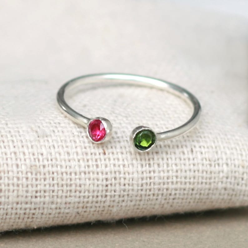 A silver cuff ring is shown from the front with pink and green gemstones adoring each end.  The ring is lying on a light-coloured resin cloth.