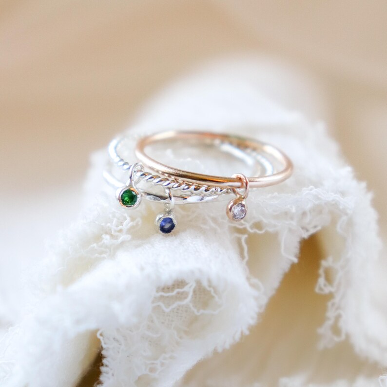 K&ouml;nnte beinhalten: Drei zarte Ringe mit baumelnden Anh&auml;ngern. Ein Ring ist ros&eacute;goldfarben, die anderen beiden sind silberfarben. Jeder Ring hat einen anderen Edelstein: gr&uuml;n, blau und klar. Die Ringe werden auf einem wei&szlig;en, strukturierten Stoff pr&auml;sentiert.