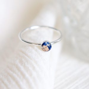 May include: A thin silver-toned metal ring featuring a small, circular bezel setting. The center of the setting contains a decorative inlay with a mix of deep blue and metallic gold-colored flecks, creating a speckled, textured appearance.