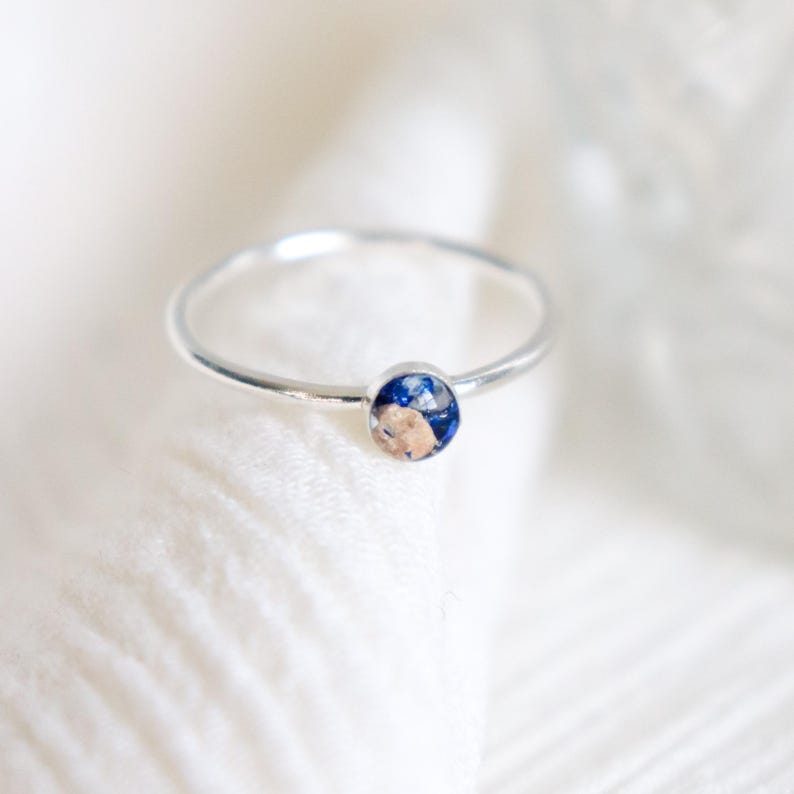 May include: A thin silver-toned metal ring featuring a small, circular bezel setting. The center of the setting contains a decorative inlay with a mix of deep blue and metallic gold-colored flecks, creating a speckled, textured appearance.