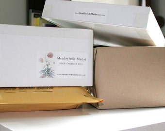 May include: Three white boxes with labels that say "Meadowbelle Market" and "Made Fresh For You!"  The boxes are stacked on top of each other on a white surface. The top box has a website address on the label. The bottom box is covered with a brown paper envelope.