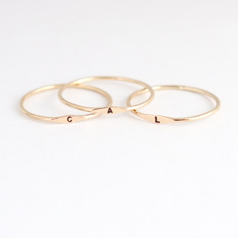 Three 1 mm handmade initial ring bands in 14k gold are neatly arranged in an overlapping stack featuring tiny letters C, A and L respectively from left to right. The rings sit on a very light off white background.